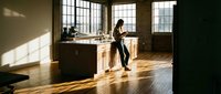 Young woman with dark hair and medium skin tone working in modern loft kitchen with wooden floors, industrial windows, and natural sunlight. Suitable for lifestyle, business, and home design projects.