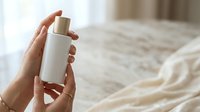 Caucasian woman's hands holding white cylindrical cosmetic bottle with gold cap against blurred bedroom background for skincare product marketing.