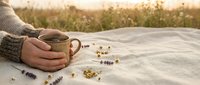 Woman in cozy sweater holding ceramic mug with herbal tea on blanket outdoors during golden hour sunset with dried flowers scattered around peaceful moment.