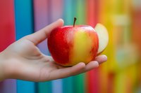 Fresh red apple held in female hand against colorful rainbow striped background, soft focus photography with vibrant colors and natural lighting.