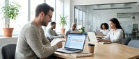 Young professional businessman with glasses working on laptop in modern office conference room with colleagues in background.
