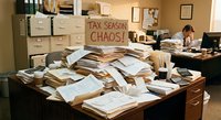 Overwhelmed office worker surrounded by massive piles of paperwork and documents during busy tax season with humorous chaos sign on wall.