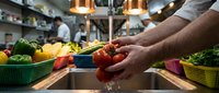 Male chef with dark skin tone washing fresh red tomatoes under running water at commercial kitchen sink, surrounded by colorful produce in baskets, preparing ingredients for meal preparation.