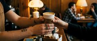 Barista handing takeaway coffee cup to customer in warm cafe interior with blurred patrons and ambient lighting in background.