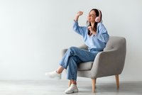 Young woman enjoying music with white headphones while sitting in modern gray armchair, wearing casual blue outfit and expressing joy through dance movements.