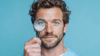 Curious man with wavy hair holding magnifying glass over eye against turquoise background, representing search, discovery, and investigation.