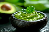 Fresh guacamole served in black avocado skin bowl garnished with basil leaves and water drops, creamy texture on dark background with blurred avocado.