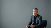 Mature male professional with gray hair and beard, wearing dark blazer and navy shirt, seated against neutral gray background for corporate portraits and business headshots.