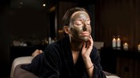 Young woman with clay mud mask on face relaxing in luxury spa salon, wearing black robe, candles glowing in background.
