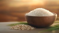 Wooden bowl filled with white rice grains beside scattered raw rice and green grass blades on wooden surface with warm golden sunset background.