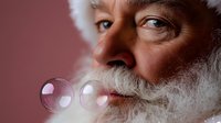 Senior man with natural white beard and kind eyes blowing soap bubbles against pink background, creating magical holiday atmosphere for seasonal greetings and celebrations.