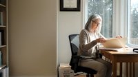 Senior Caucasian woman with gray hair and glasses working on laptop at wooden desk in bright home office, focused on computer screen for remote work.
