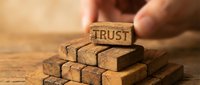 Hand placing wooden block with TRUST text on top of pyramid stack of similar blocks on wooden surface representing business foundation and reliability concepts.