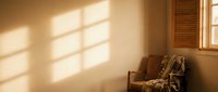 Warm sunlight streams through window casting geometric shadows on empty beige wall with wooden chair in corner, creating serene minimalist interior design atmosphere.