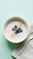 Creamy yogurt bowl topped with fresh blueberries and toasted coconut flakes, served with spoon on mint green background for breakfast and healthy eating concepts.