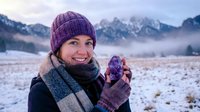 Young Caucasian woman in purple knit hat and mittens drinking hot beverage in snowy mountain landscape during winter morning with misty peaks backdrop.