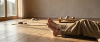 Person resting barefoot on wooden floor in a serene zen meditation room with natural light, stones, and tea set in background.