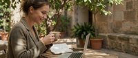 Young professional woman enjoying coffee while working on laptop in sunny outdoor cafe setting with natural stone wall and green plants creating peaceful workspace.
