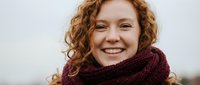Young woman with curly red hair wearing burgundy knitted scarf smiling warmly at camera against neutral background for lifestyle and fashion content.