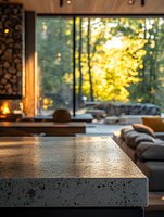 Modern minimalist living room with concrete coffee table and large windows overlooking lush green forest creating warm natural lighting atmosphere.