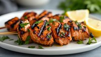 Grilled shrimp skewers with charred edges, garnished with fresh herbs and lemon wedges on white plate. Closeup of juicy seafood appetizer showing caramelized glaze.