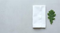 Blank white napkin or paper towel next to fresh green oak leaf on neutral gray surface for design mockups and branding presentations.