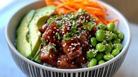 Fresh poke bowl with raw tuna, avocado slices, edamame beans, shredded carrots and sesame seeds served in ceramic bowl on wooden surface.