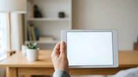 Hand holding tablet with blank white screen in home office workspace with wooden desk, shelving, and plant.