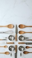 Kitchen utensils and cookware arranged symmetrically on white marble surface including wooden spoons, measuring cups, whisk and knife for cooking.
