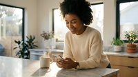 Young woman of African descent with curly hair wearing a beige sweater, smiling while using a smartphone at a wooden table in a bright, plant-filled home office space.