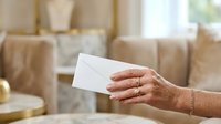 Woman's hand holding blank white envelope in modern living room with neutral furnishings, suitable for business correspondence, invitations, and greeting card concepts.