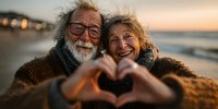Happy senior couple making heart shape with hands at sunset beach, romantic elderly man and woman enjoying golden hour together by ocean pier.