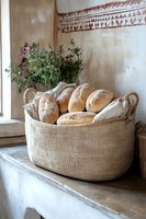 Fresh baked bread loaves in rustic linen basket on windowsill with herbs, natural light setting creates cozy farmhouse atmosphere for food photography.