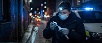 Male detective examining evidence with flashlight on urban street at night wearing protective mask and gloves during investigation.