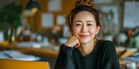 Young Asian woman with messy bun hairstyle smiling warmly in cozy cafe, resting chin on hand, wearing dark sweater against blurred interior background.