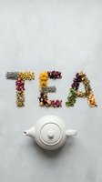Word TEA spelled with colorful dried herbs, flowers, and leaves beside a white ceramic teapot on a light concrete surface.