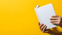 Person with medium skin tone holding open blank white book against bright yellow background, suitable for education and publishing concepts.