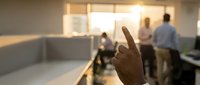 Close-up of a hand with index finger pointing in a modern office workspace with blurred colleagues working in the background.