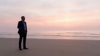 Silhouette of businessman in suit standing on beach at sunset contemplating future opportunities and career success with ocean waves and pink sky.
