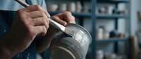 Skilled artisan hands carefully painting decorative details on ceramic pottery piece in traditional workshop studio setting for handmade crafts and artistic creation.
