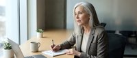 Mature businesswoman with gray hair working on laptop at office desk, professional female executive in suit planning strategy during meeting preparation.