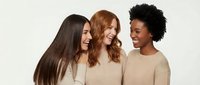 Three diverse women with different hair textures laughing together in matching beige sweaters against neutral background for lifestyle campaigns.