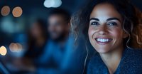 Young mixed race woman with curly hair smiling warmly at night, with soft bokeh lights and blurred people in background creating atmospheric mood.