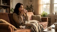 Young woman with long dark hair and olive complexion sitting on beige sofa in warm living room, holding tissue, appearing contemplative or unwell. Suitable for healthcare, wellness, mental health and lifestyle editorial use.