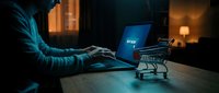 Young man shopping online at night, typing on laptop with buy now button on screen and mini cart on desk in dark room.