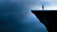 Silhouette of person standing on edge of dramatic cliff against moody blue sky, representing risk, challenge, courage and achievement in business concept.