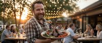 Smiling middle-aged Caucasian male chef in plaid shirt serving grilled meat platter at outdoor restaurant during golden hour sunset dining experience.