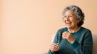Senior woman with gray hair, Caucasian ethnicity, wearing teal sweater, smiling warmly while holding microphone against neutral beige background for speaking engagement or presentation use.