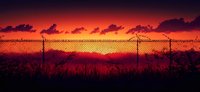 Dramatic orange sunset sky with dark clouds silhouetted behind chain link fence, creating moody atmospheric panoramic landscape with wild grass in foreground.