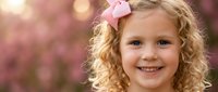 Happy blonde curly-haired girl with pink bow smiling outdoors against soft floral background. Child portrait for family photography and spring concepts.